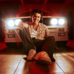 Man sitting on a garage floor in front of a white car with bright headlights, smiling at the camera under red lighting.