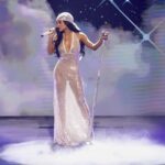 Female singer in a sparkling champagne gown performs on a foggy stage with a microphone and stand, starry backdrop.