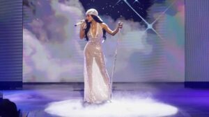 Female singer in a sparkling champagne gown performs on a foggy stage with a microphone and stand, starry backdrop.