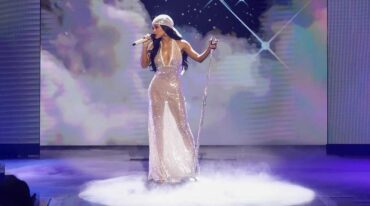 Female singer in a sparkling champagne gown performs on a foggy stage with a microphone and stand, starry backdrop.