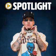 Person with multiple tattoos, wearing a cap and sunglasses, holding fists near chin against a dark blue backdrop; graphic tee visible.