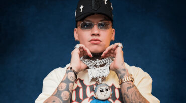Person with multiple tattoos, wearing a cap and sunglasses, holding fists near chin against a dark blue backdrop; graphic tee visible.