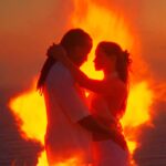 Couple embracing in white outfits with a large bonfire behind them on a beach at sunset.