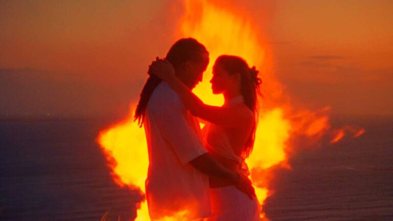 Couple embracing in white outfits with a large bonfire behind them on a beach at sunset.