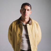 Portrait of a man in a tan jacket with a brown collar, layered necklaces, standing against a gray studio background.