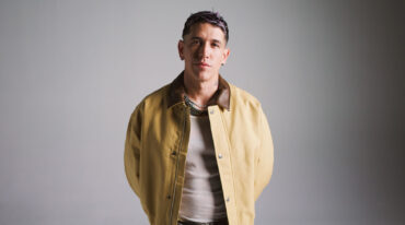 Portrait of a man in a tan jacket with a brown collar, layered necklaces, standing against a gray studio background.