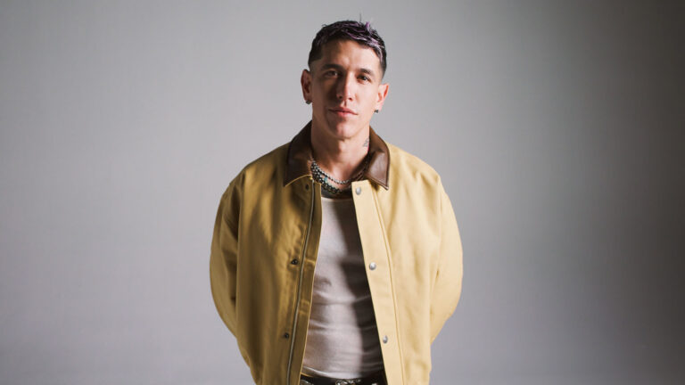 Portrait of a man in a tan jacket with a brown collar, layered necklaces, standing against a gray studio background.