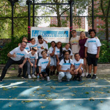 Group of children and adults posing for a photo on a blue outdoor basketball court with a banner behind them.