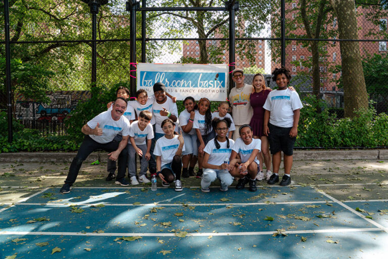 Group of children and adults posing for a photo on a blue outdoor basketball court with a banner behind them.