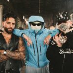 Three men pose in a gritty industrial space: a tattooed man in a black leather vest, a blue-jacketed figure with a balaclava and sunglasses, and a heavily tattooed man with a cap and chain—center foreground.
