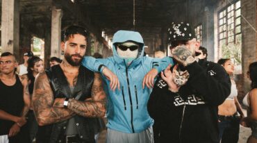 Three men pose in a gritty industrial space: a tattooed man in a black leather vest, a blue-jacketed figure with a balaclava and sunglasses, and a heavily tattooed man with a cap and chain—center foreground.