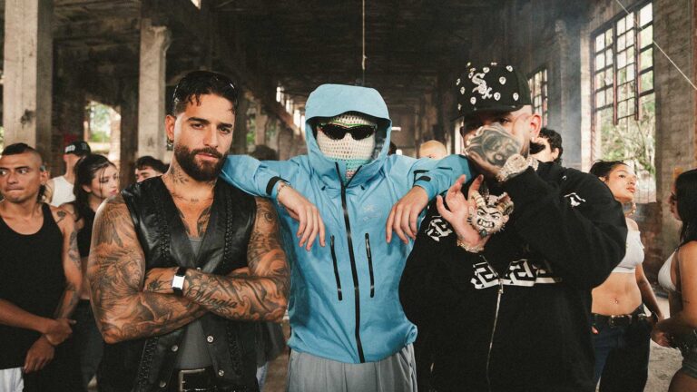 Three men pose in a gritty industrial space: a tattooed man in a black leather vest, a blue-jacketed figure with a balaclava and sunglasses, and a heavily tattooed man with a cap and chain—center foreground.