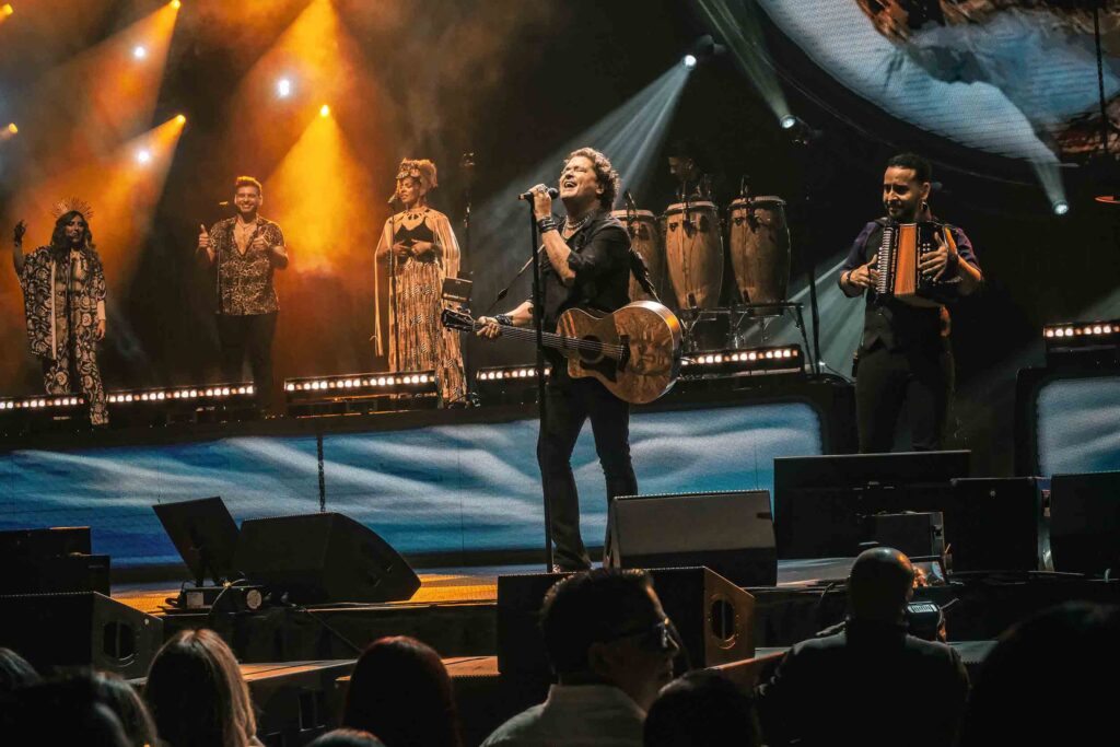Live concert scene with a five-member band performing on a lit stage; center singer with acoustic guitar, surrounded by other vocalists/instrumentalists under warm orange lights, with percussion in the back and an accordion player on the right.