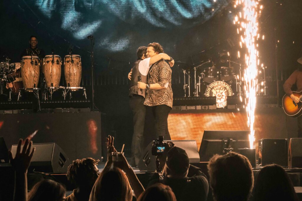 Two performers embrace on a concert stage as fireworks shoot overhead, with a drummer on the left and cheering audience in front.
