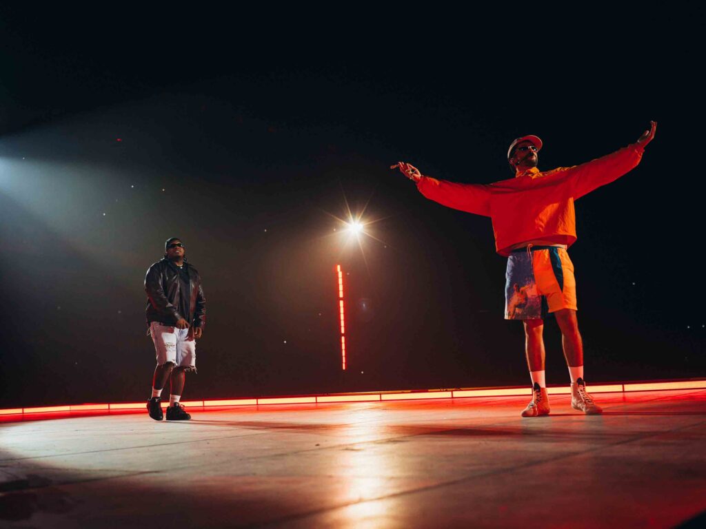 Two performers on a dark stage; one in bright orange with arms raised, the other in a black jacket watching nearby under stage lights.
