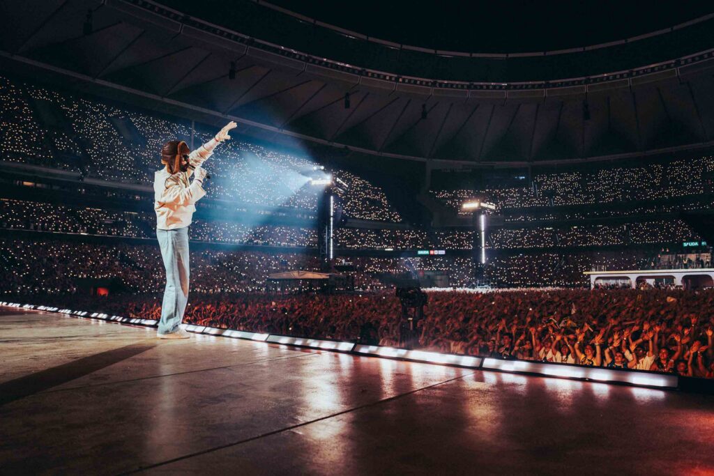 Performer on a large stadium stage raises an arm toward the cheering crowd, thousands of tiny lights filling the audience.
