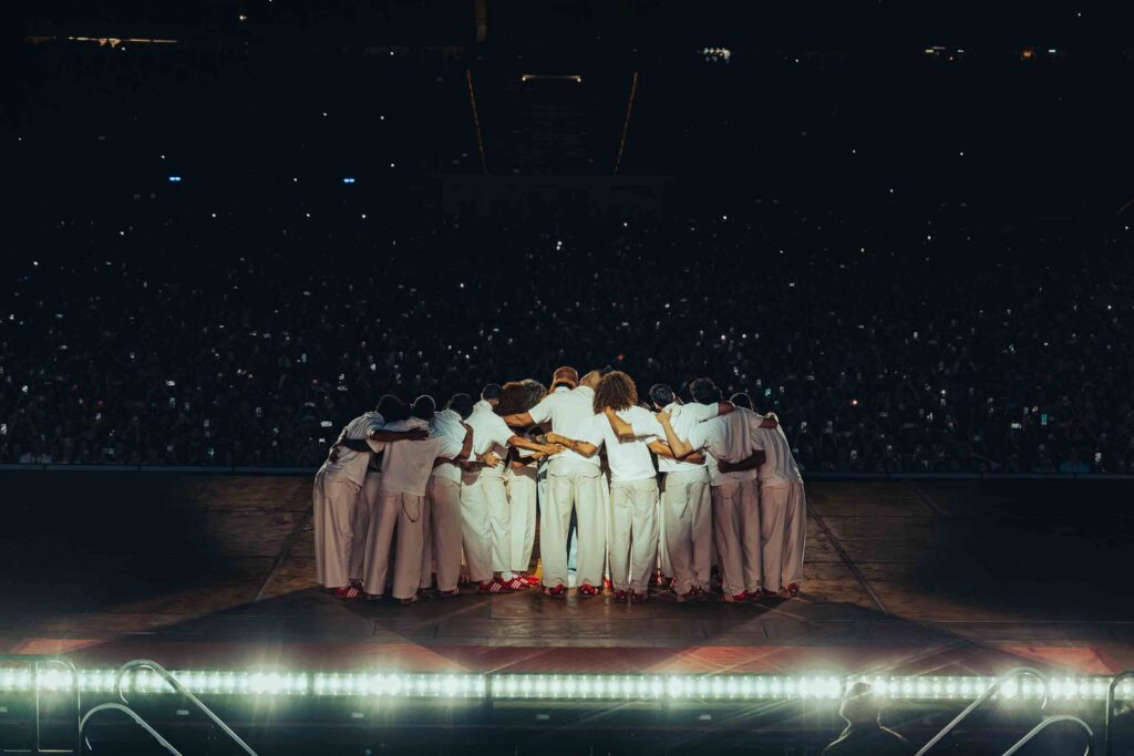 Group of performers in white outfits with arms around each other, forming a circle on a stage before a large nighttime audience.