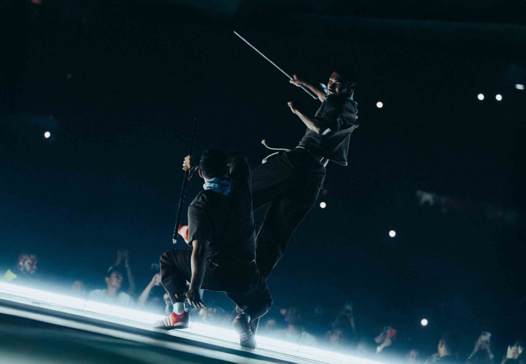 Two performers balance on a glass stage in a dark arena, holding long poles as bright lights twinkle in the background.]
