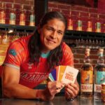 Person in red shirt at a bar holding colorful cards, with tequila bottles and a decorated cake nearby in the foreground.