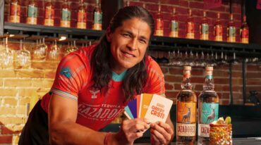 Person in red shirt at a bar holding colorful cards, with tequila bottles and a decorated cake nearby in the foreground.