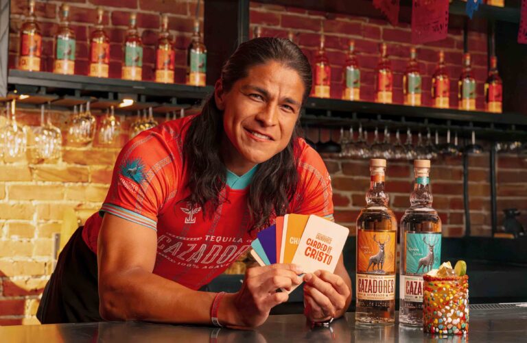 Person in red shirt at a bar holding colorful cards, with tequila bottles and a decorated cake nearby in the foreground.