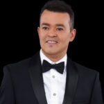 Portrait of a man in a black tuxedo and bow tie against a black background, smiling slightly.