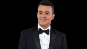 Portrait of a man in a black tuxedo and bow tie against a black background, smiling slightly.