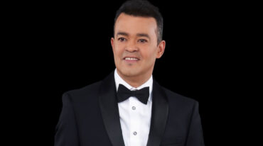Portrait of a man in a black tuxedo and bow tie against a black background, smiling slightly.