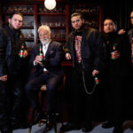 Older man in a suit seated on a bar stool, surrounded by six men in black leather jackets, all holding beer bottles in a dim bar setting.