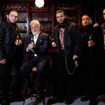 Older man in a suit seated on a bar stool, surrounded by six men in black leather jackets, all holding beer bottles in a dim bar setting.
