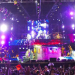 Crowd with raised hands watching a large, multicolored concert stage and band performing under bright lights.