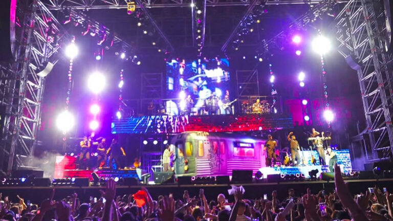 Crowd with raised hands watching a large, multicolored concert stage and band performing under bright lights.
