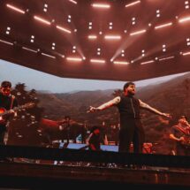 Live concert scene with a guitarist and a vocalist on a lit stage; large video backdrop shows a desert landscape under red lights.