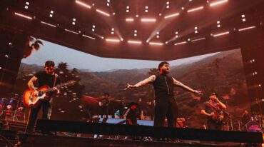 Live concert scene with a guitarist and a vocalist on a lit stage; large video backdrop shows a desert landscape under red lights.