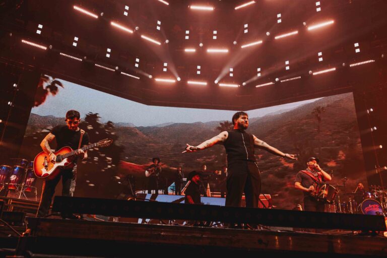 Live concert scene with a guitarist and a vocalist on a lit stage; large video backdrop shows a desert landscape under red lights.