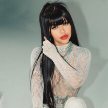 Young woman with long black hair wearing a white lace long-sleeve outfit and chunky silver necklaces posing against a pale backdrop.