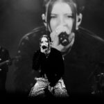 Lead female singer at center stage, microphone in hand, Leopard-print pants, band and large screen backdrop in grayscale photo.