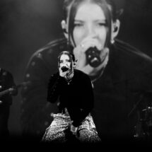 Lead female singer at center stage, microphone in hand, Leopard-print pants, band and large screen backdrop in grayscale photo.