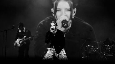 Lead female singer at center stage, microphone in hand, Leopard-print pants, band and large screen backdrop in grayscale photo.