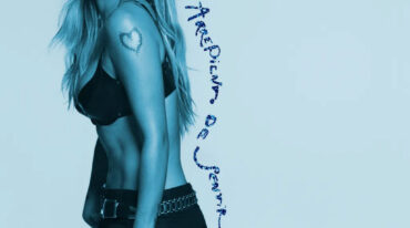 Portrait of a woman in a black bra and pants, shown in a side profile, with a heart tattoo on her upper arm and a blue vertical handwritten caption along her side.